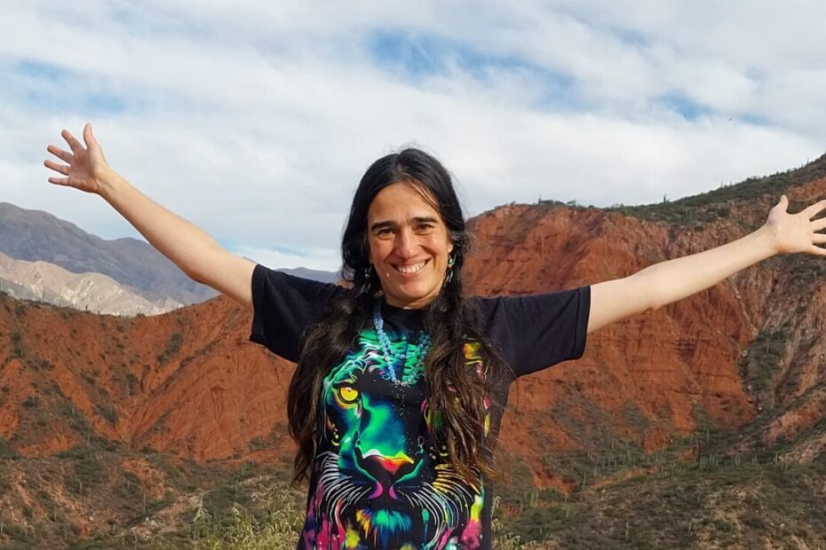 Cata Heincke con brazos abiertos en un paisaje de montañas rojas, ofreciendo mentoría 1 a 1 online personalizada.