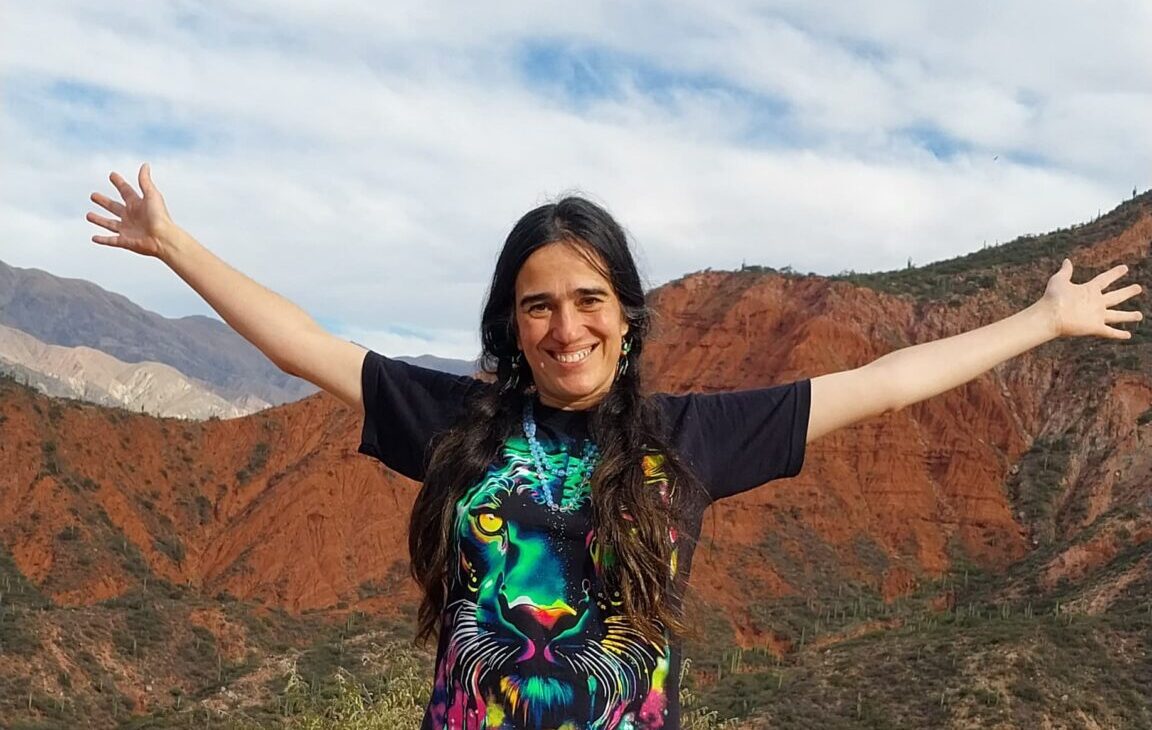 Cata Heincke con brazos abiertos en un paisaje de montañas rojas, ofreciendo mentoría 1 a 1 online personalizada.